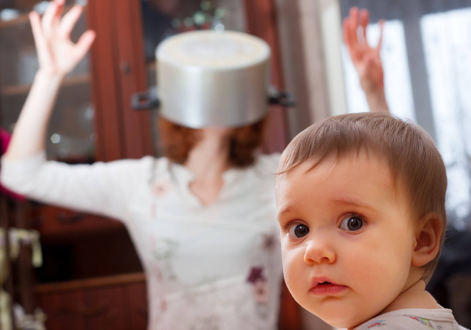 Toddler looks at us as mother is stressed in the background with a pot over her head.